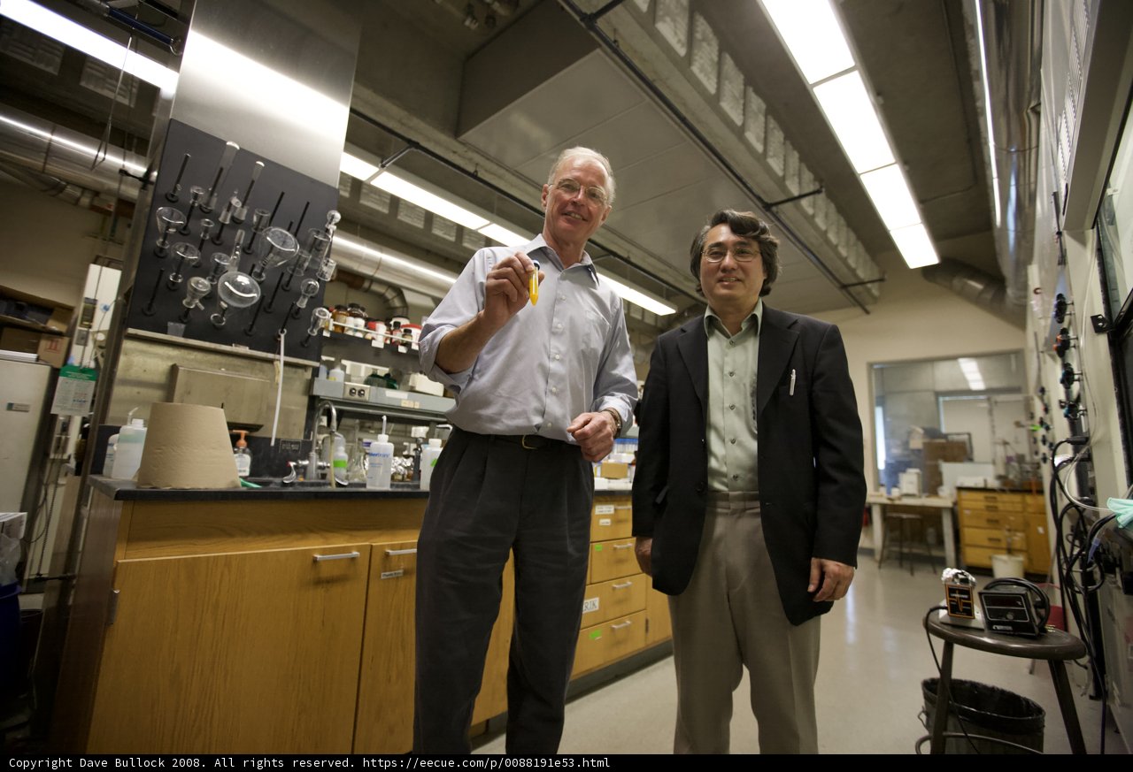 Two Men Working in a Nanomachine Laboratory Caption: Two men dressed in suits and coats stand in a laboratory, analyzing a glass sample under the bright lights. Plywood shelves and furniture line the walls, displaying various accessories and glassware.