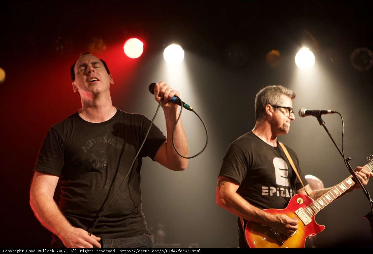 Rocking the Stage Caption: Two male musicians play electric guitars and sing into microphones while lit by a spotlight during a crowd-thrilling performance at the 2007 Bad Religion Glasshouse concert.