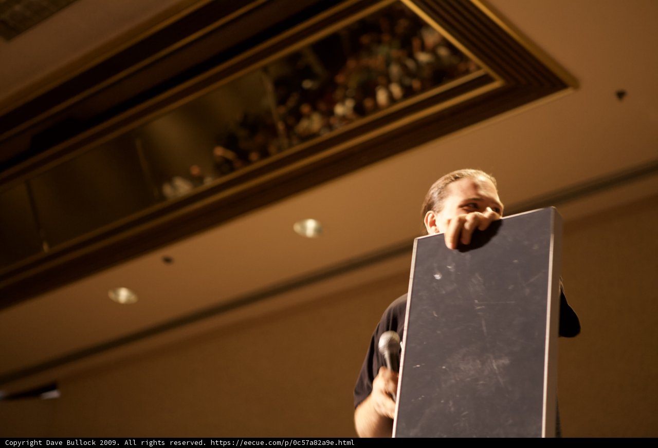 Speaking to the Crowd Caption: A man delivers a lecture holding a large blackboard while standing in front of a room filled with people and electronics.