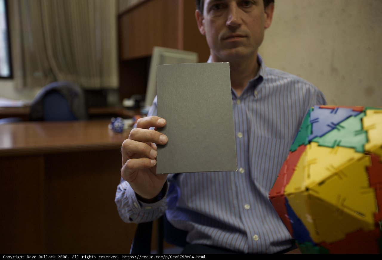 Man holding an important paper Caption: A man with a finger holding a piece of paper while sitting on a wooden chair in an indoor setting with a laptop and some boxes on the table