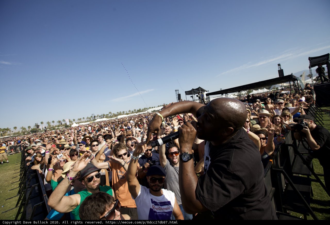Shōgo Hamada Rocks Coachella Caption: Shōgo Hamada stands proud in front of a lively crowd at Coachella 2010. His all-black clothing and sunglasses add an aura of coolness to the concert atmosphere. The blue sky and fence surrounding the land add to the outdoor recreation vibe.