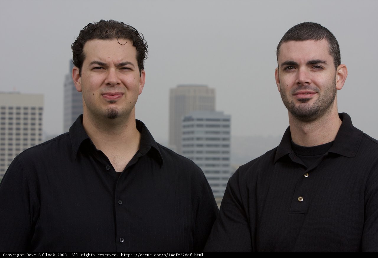 Two Men in Black Shirts Smile in Front of Skyscraper