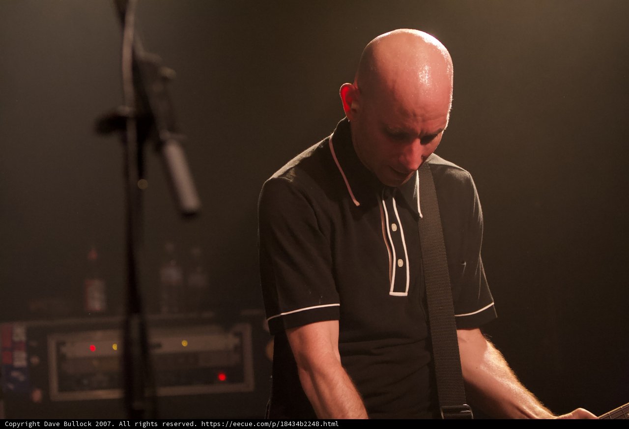 Rocking Out on Stage Caption: A bald man jams on his guitar in front of a microphone during a Bad Religion Glasshouse concert in 2007. The crowd cheers him on as he performs an electrifying solo for the audience.