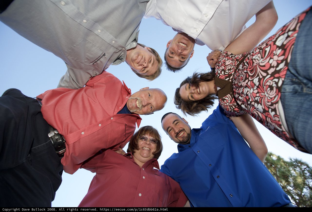 Circle of Friends Caption: Eight people, three women and four men, wearing a mix of casual and formal clothing, huddle together under a bright blue sky.