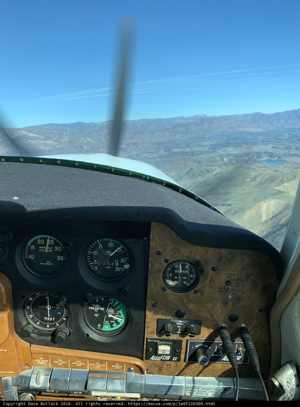 High in the Sky Caption: A close-up view of the cockpit dashboard and tachometer of a small airplane soaring over hills and land.