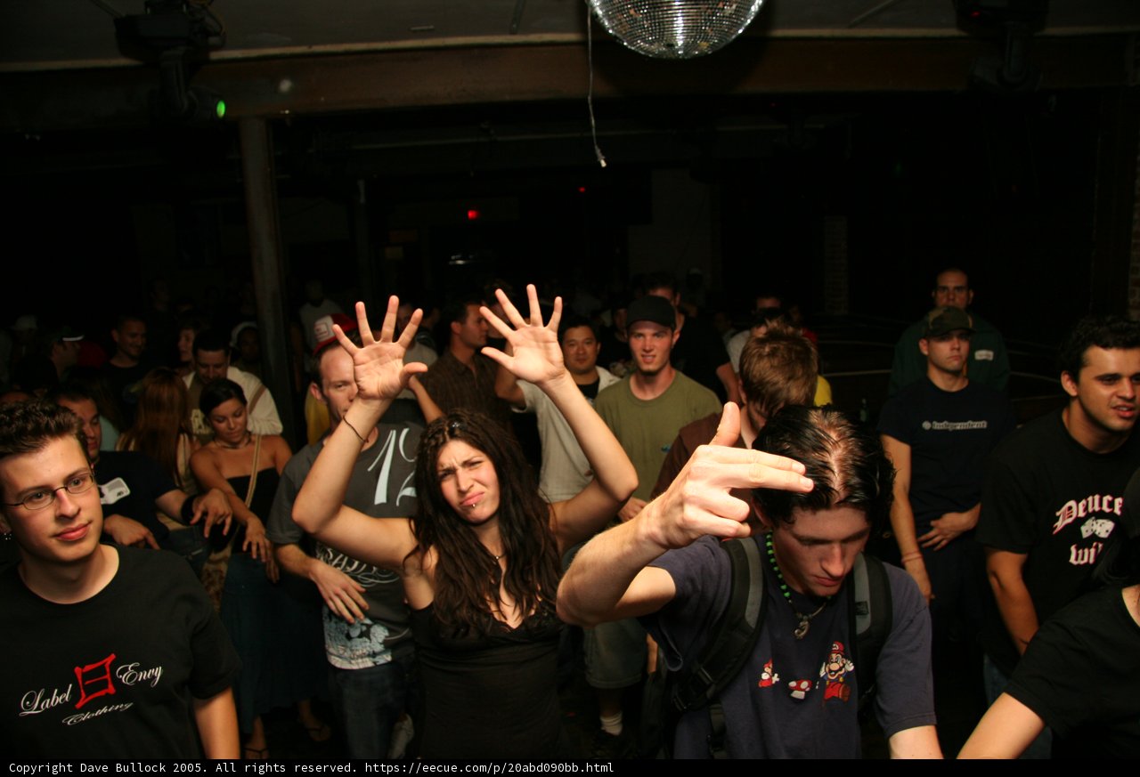 Black-Shirted Man in the Crowd Caption: This man stands out in the midst of a crowded nightclub, wearing a black shirt and accessorized with two necklaces and glasses.