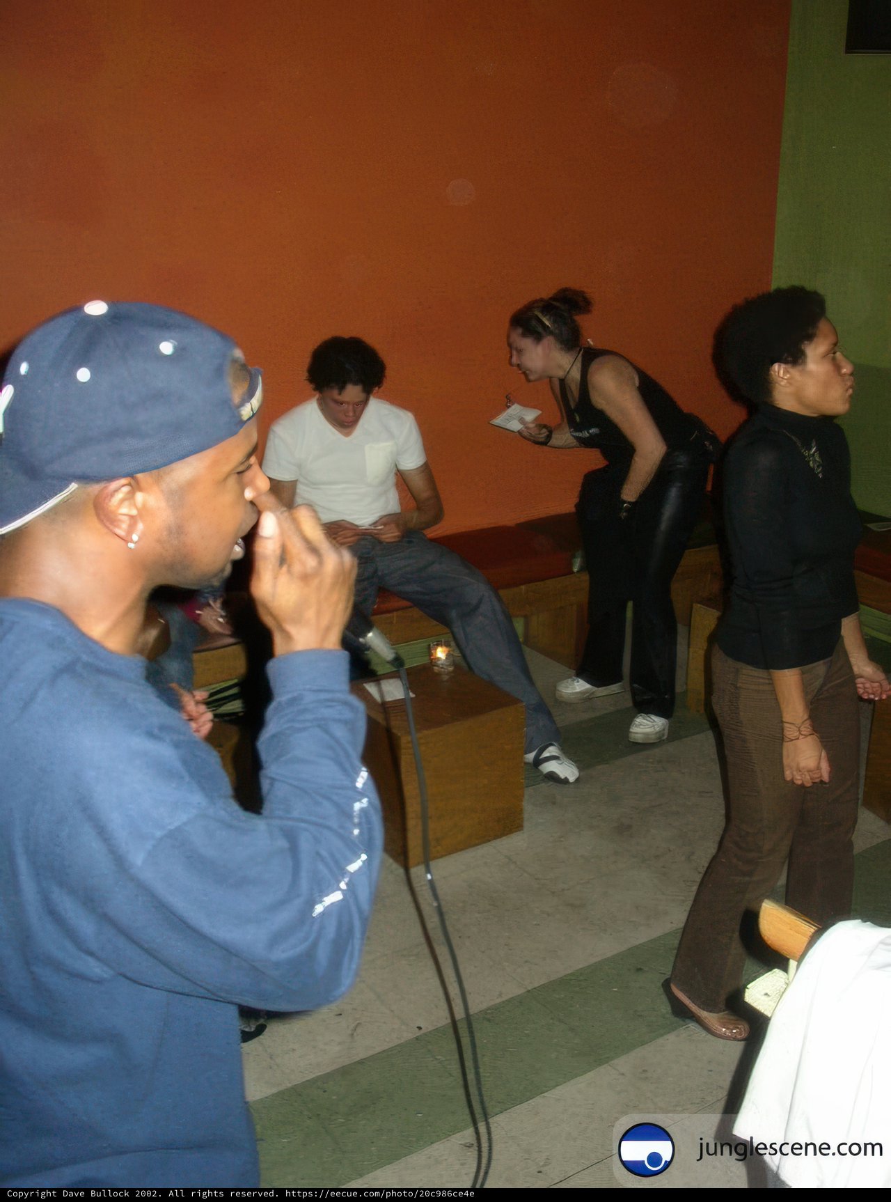 Man Holding Microphone Caption: A man wearing a baseball cap holds a microphone while sitting on a couch surrounded by friends wearing bracelets, necklaces, and other accessories.
