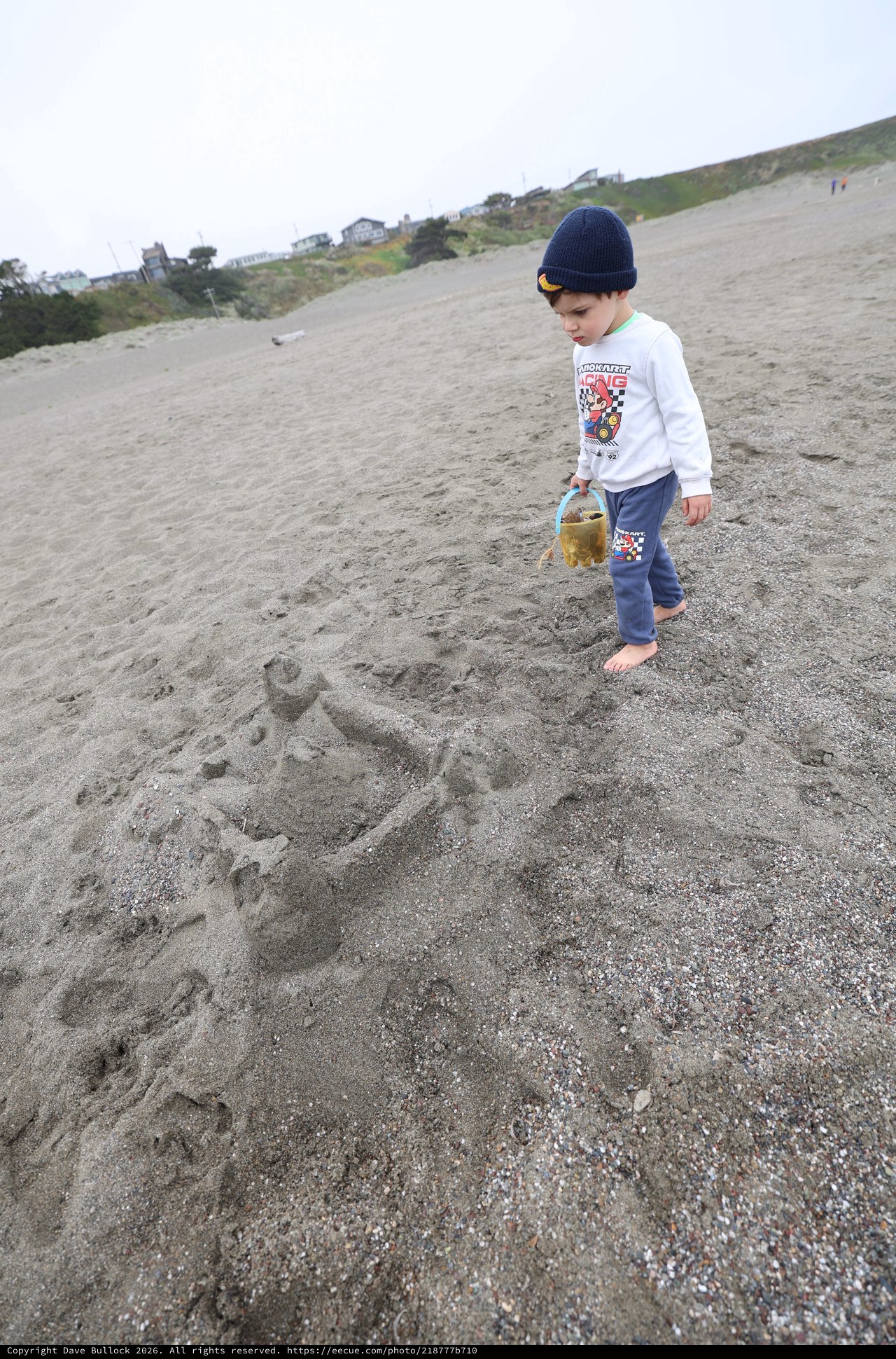 Young Boy Explores Gray Sand Beach with Bucket