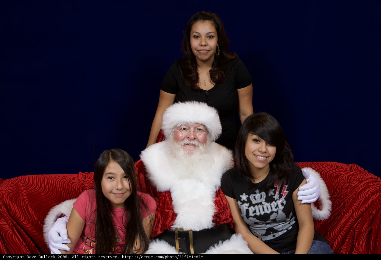 Santa Claus and the Merry Group Caption: Santa Claus poses with two girls and a woman at the 2008 APC Christmas party, all gathered comfortably on a couch.