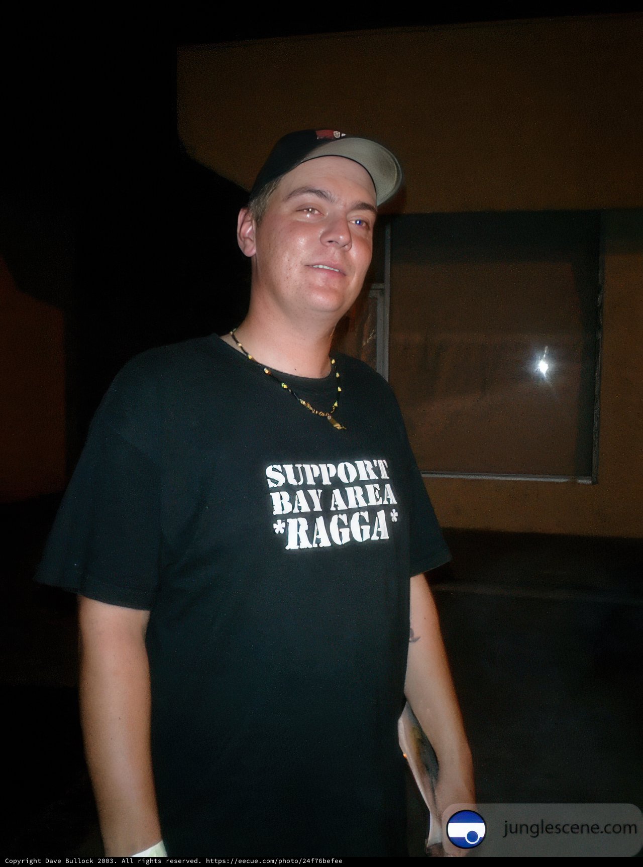 Supporting Mike Taylor Caption: A portrait of a man wearing a black shirt and baseball hat with the words "Support Mike Taylor" while flaunting his accessories and a calm demeanor in nature.