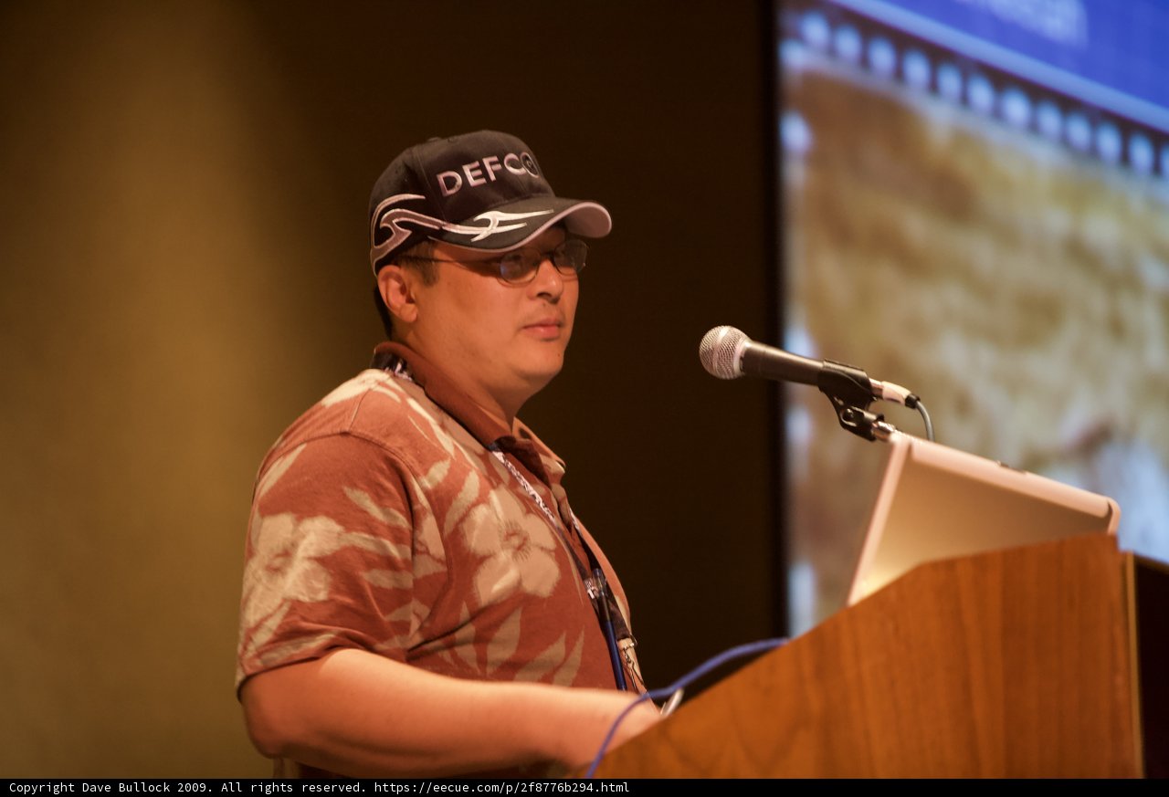 Yoshitomo Tani addresses the crowd Caption: Donning a baseball cap and sunglasses, Yoshitomo Tani delivers a speech at Defcon 17 while standing at the podium and speaking into a microphone.