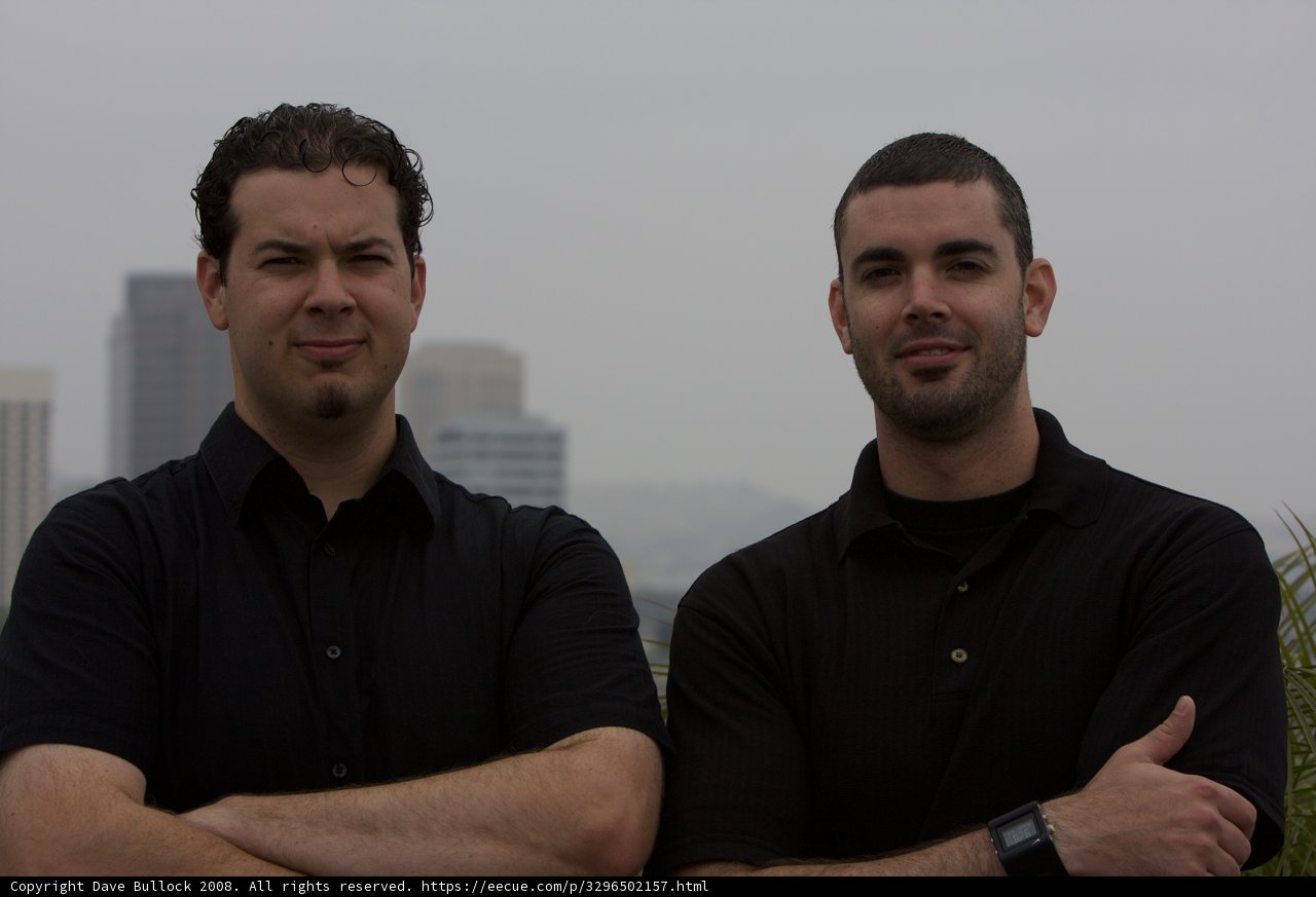 Two Men in Black Shirts Pose in Hazy Outdoor Scene