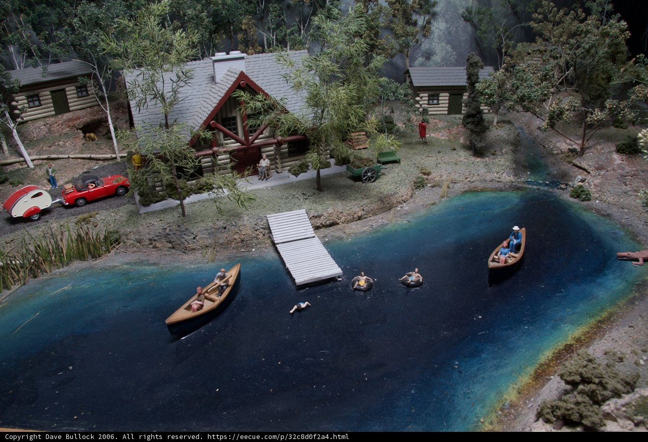 Lakeside House and Boats Caption: A miniature model of a cozy lakeside house with two boats docked in the water, surrounded by lush vegetation and bustling with activity with nine people enjoying various water sports and boat rides.