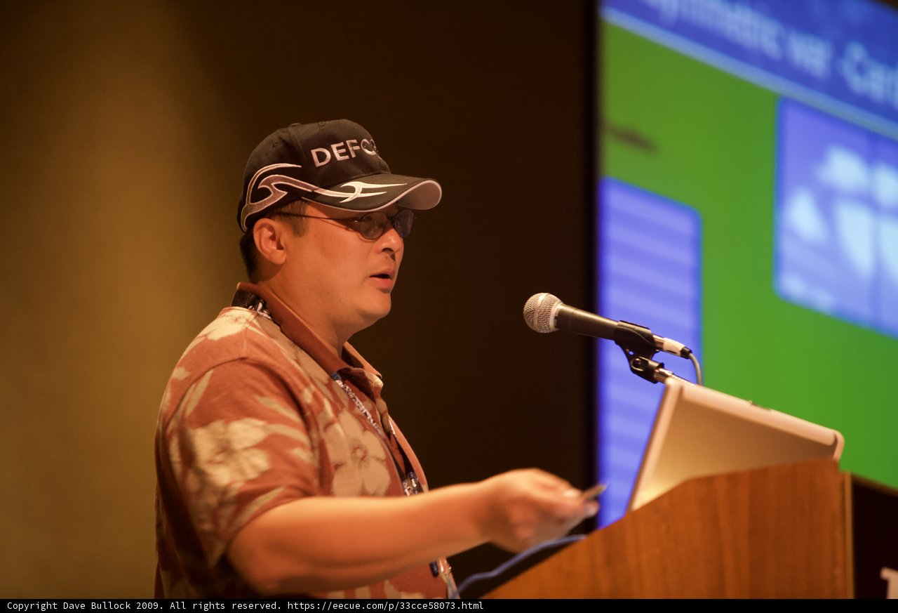 Man addresses a crowded room wearing a hat and holding a microphone