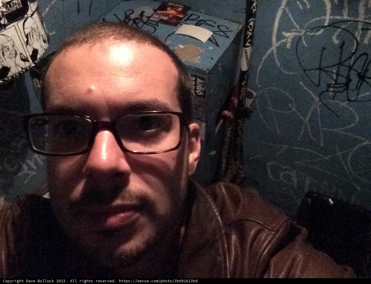 Self-portrait of a Man with Glasses and Brown Jacket in Graffiti-Filled Bathroom