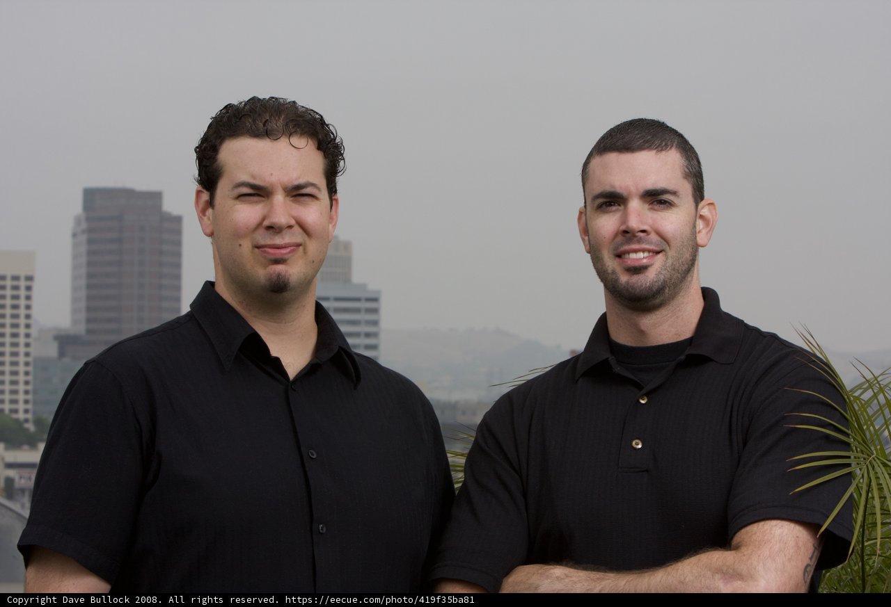 Two Smiling Men in Black Shirts with a Skyscraper in the Background