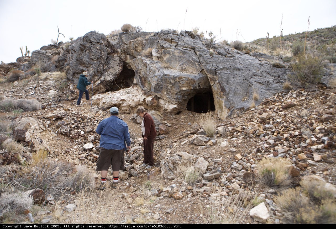 Exploring the Wilderness Caption: Two adventurers stand in front of a cave, taking shelter amongst the rocks and shrubs of the rugged outdoors.