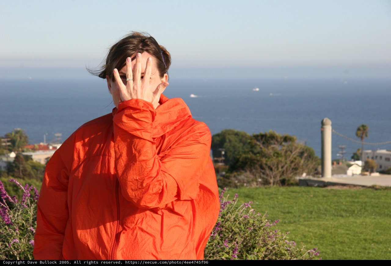 Woman in Orange Jacket Shields Face by the Sea