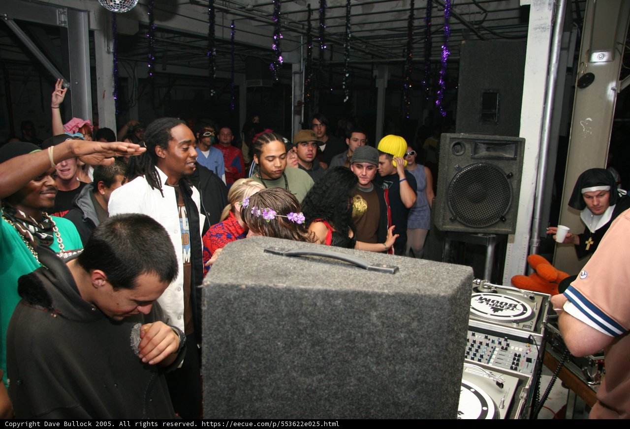 Nightclub Groove Caption: A deejay entertains a crowd of 24 people with music while standing beside the mixer and speakers. Three women and five men dance while others enjoy the performance inside the club.