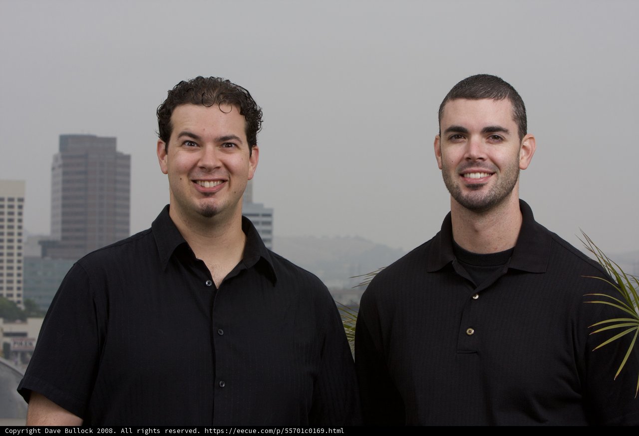 Two Smiling Men in Black Shirts Pose in Front of Cityscape