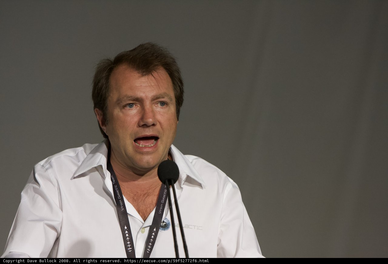 The White Knight Two Speaks Caption: A man in a white shirt addresses the crowd with a microphone in hand, commanding attention in his lab coat and dress shirt.