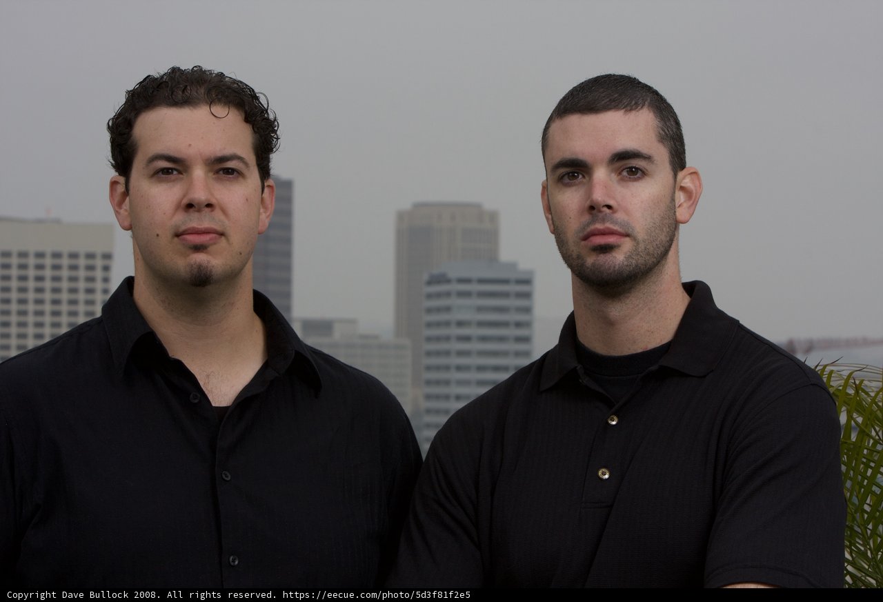 Two Men in Black Shirts Posing in Front of Skyscraper
