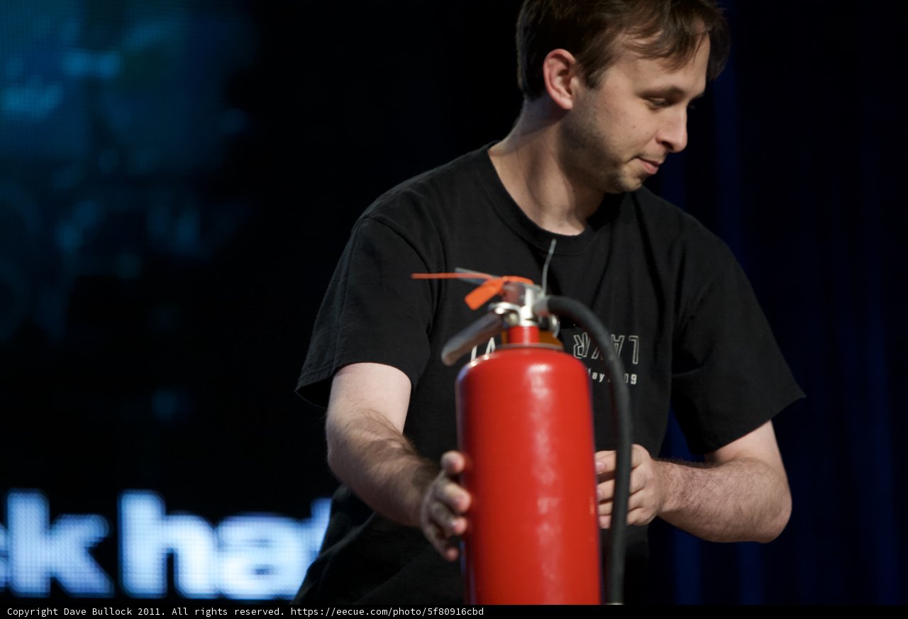 Ready to Fight Caption: A man stands with a fire extinguisher in hand, poised to battle any potential flames as he faces a screen full of people.