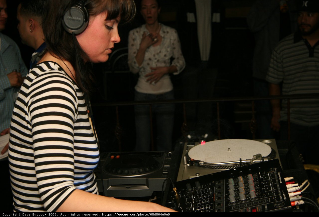 DJ Entertaining the Crowd Caption: A female deejay wearing headphones and standing behind a turntable entertains the crowd with her music at a club.