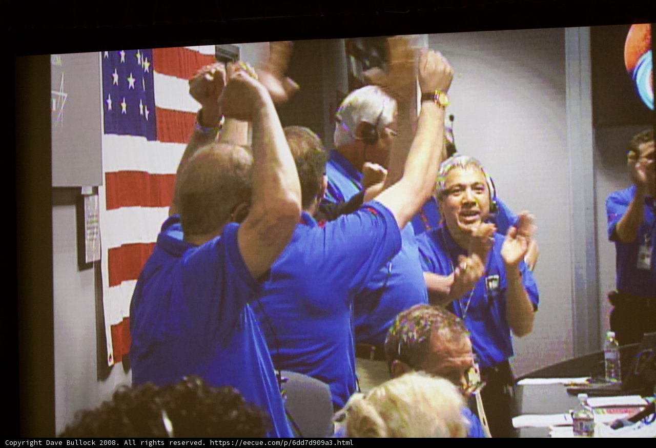 Celebrating Victory Caption: A group of 5 men wearing blue shirts cheer excitedly in front of a television displaying a football game at a bar, as one man holds up a flag in celebration.