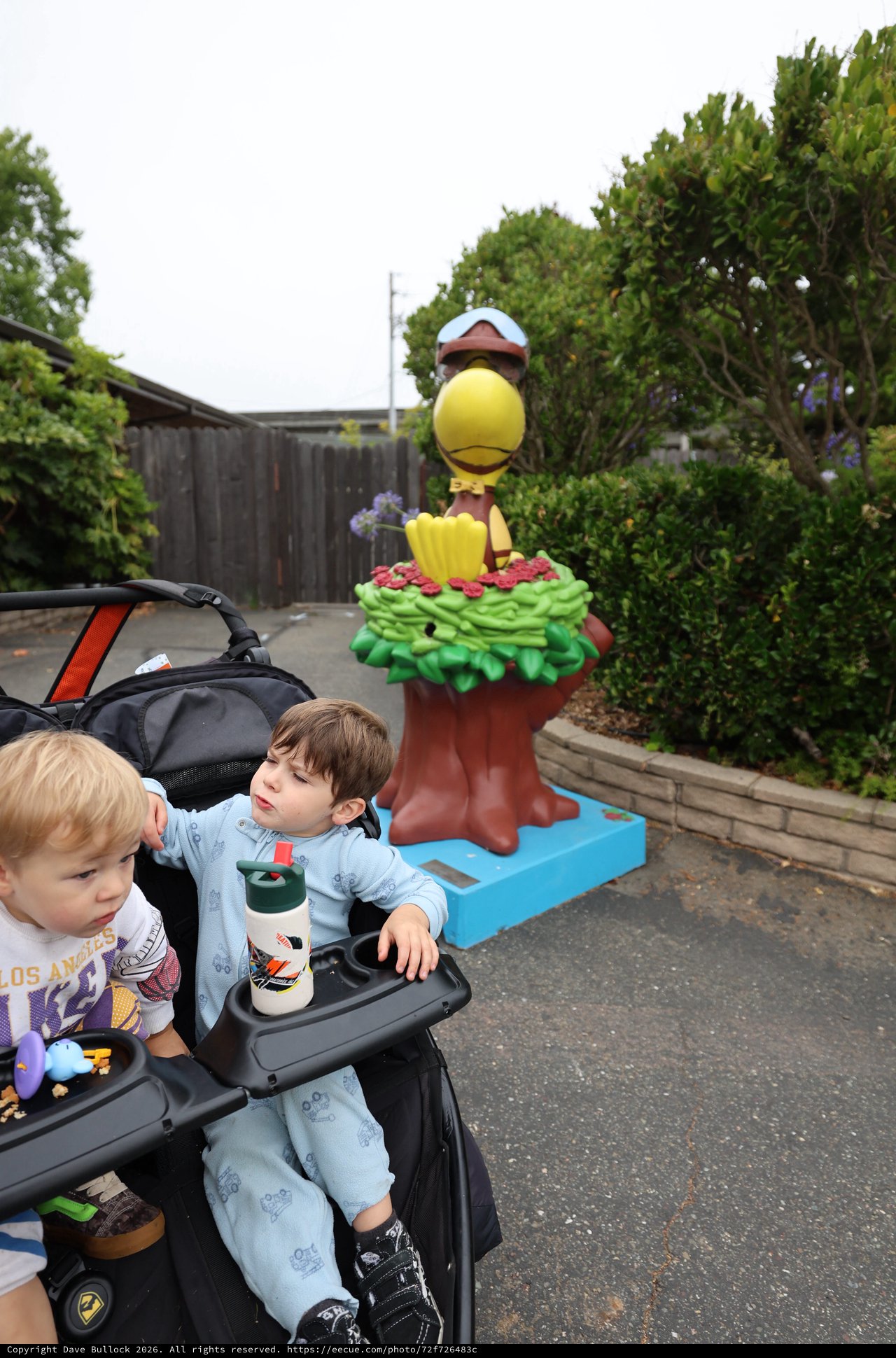 Two Young Boys in Stroller Near Cartoon Bird Statue