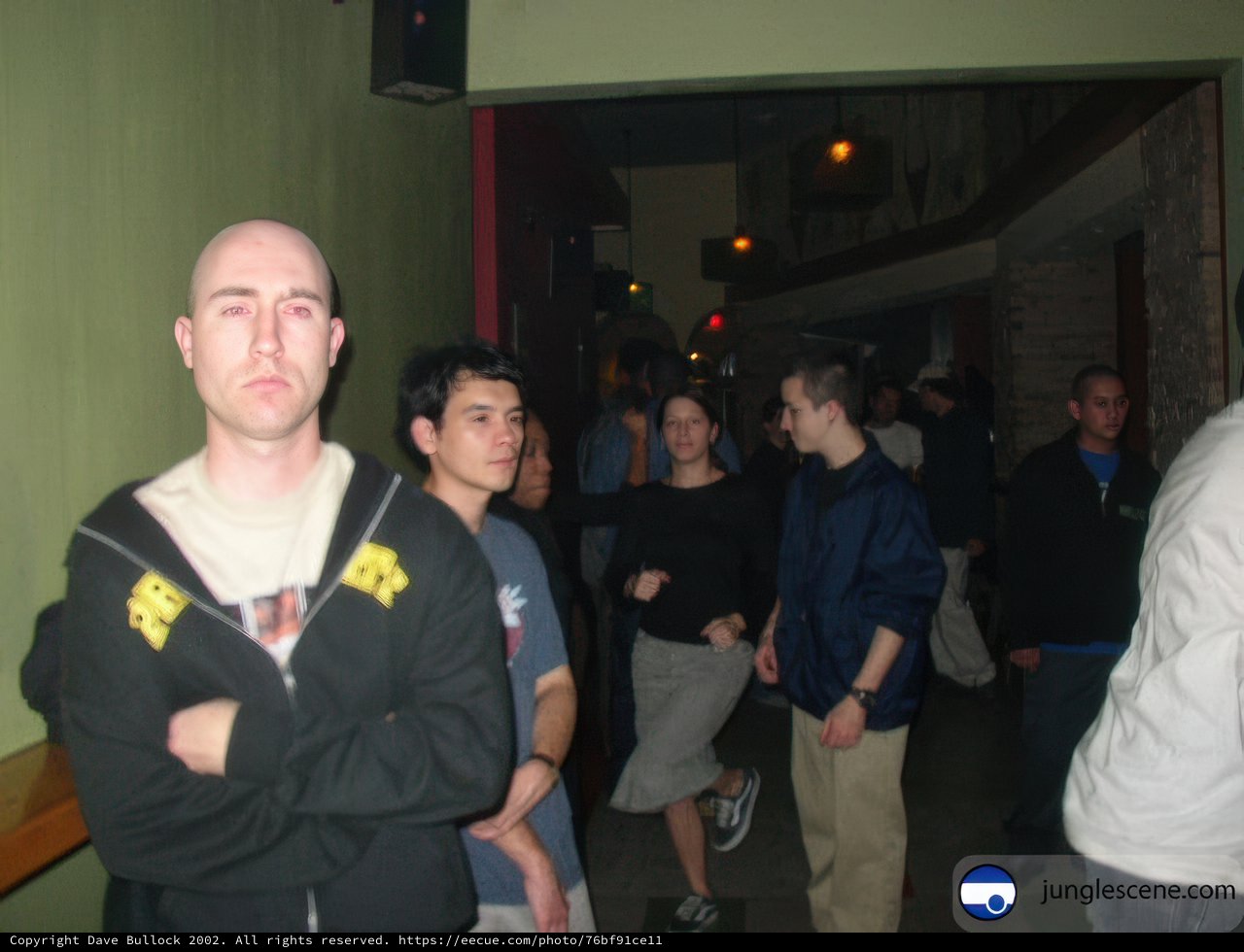 Cool and Composed Caption: Eric W stands confidently in the hallway, his arms crossed, donning a stylish jacket and footwear, ready to take on the music scene with his friend Bruce K.