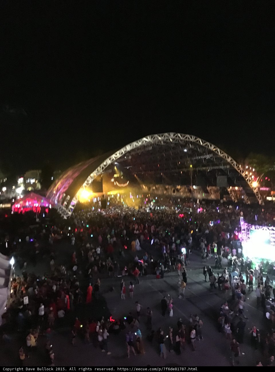 Nighttime Metropolis Crowd Caption: A large group of people gather in the heart of San Bernardino, surrounded by the bright lights of the city and the night sky, for a concert event.