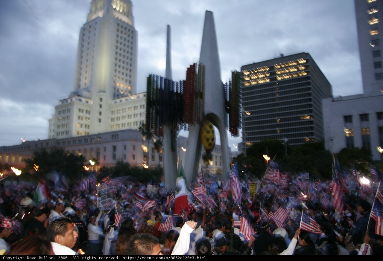 Metropolis Unites Caption: A crowd of 13 people holding flags gather in front of an office building in the city during a cloudy day.