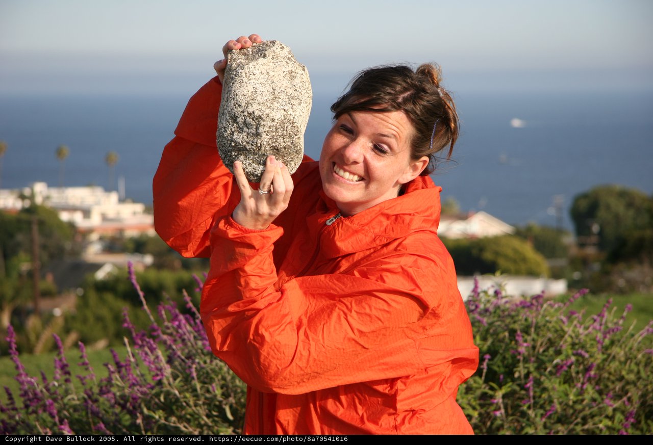 Woman Playfully Holds Giant Rock by the Sea