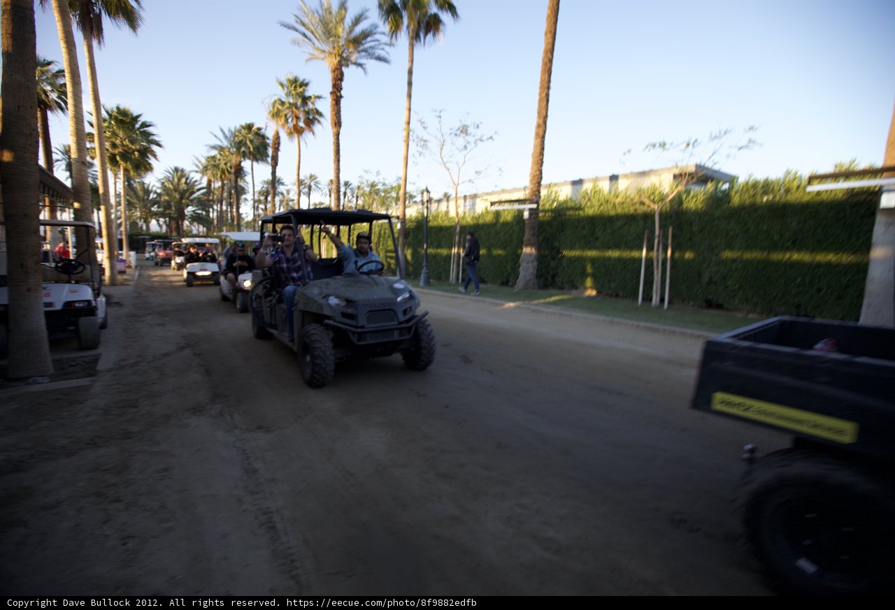Coachella Cruise Caption: Nine festival goers take a joy ride on a golf cart down a rustic road, taking in the sights of palm trees and shrubs amidst a blue sky.