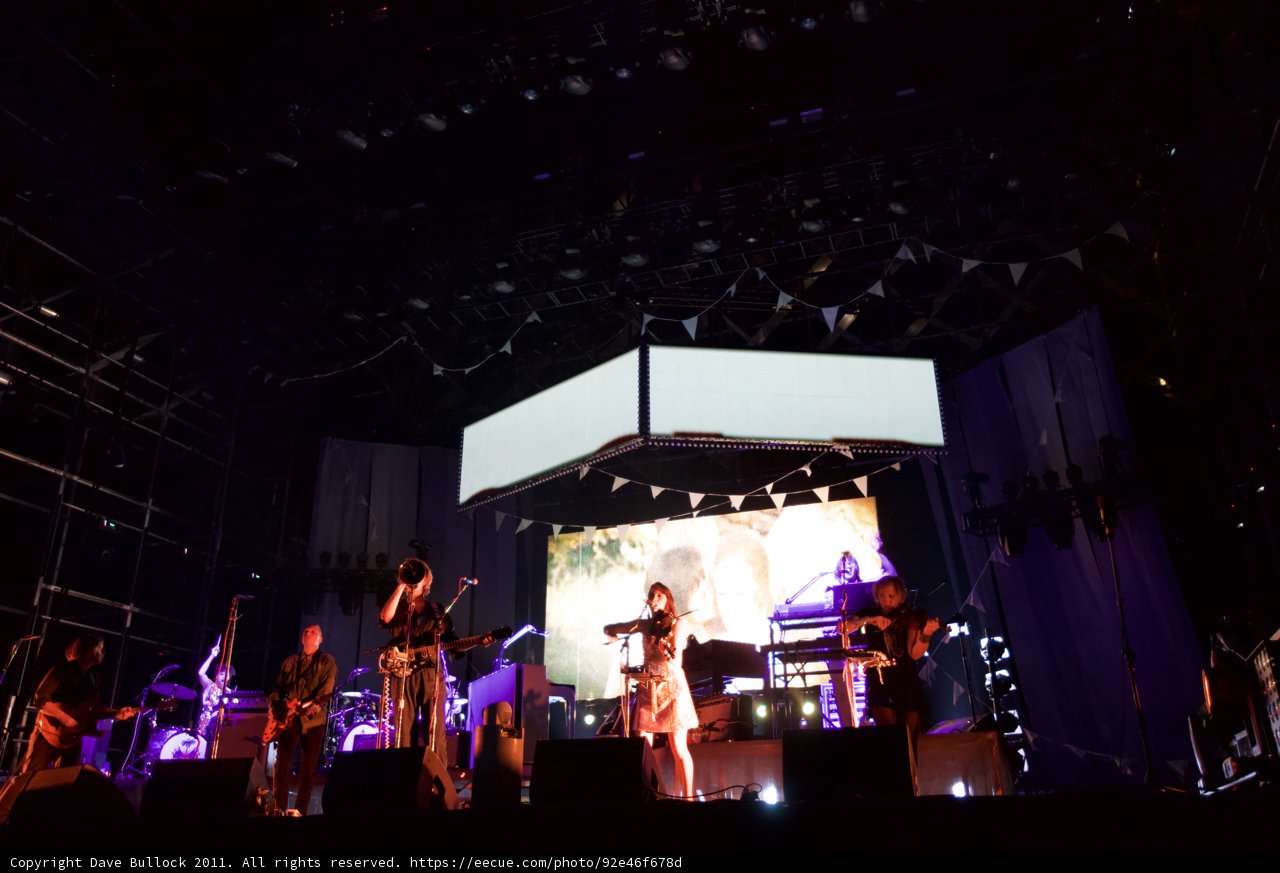 Saturday Night Rockin' at Coachella 2011 Caption: A crowd of 9 people gathered to hear the urban music of a musical instrument and three guitars at a performance by a lighting-wrapped band on stage. The electronics and seven speakers added to the enthralling concert experience.