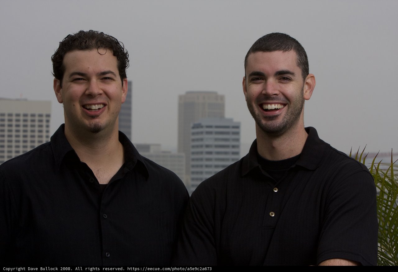 Smiling Duo in Black Shirts Pose Against a Skyscraper