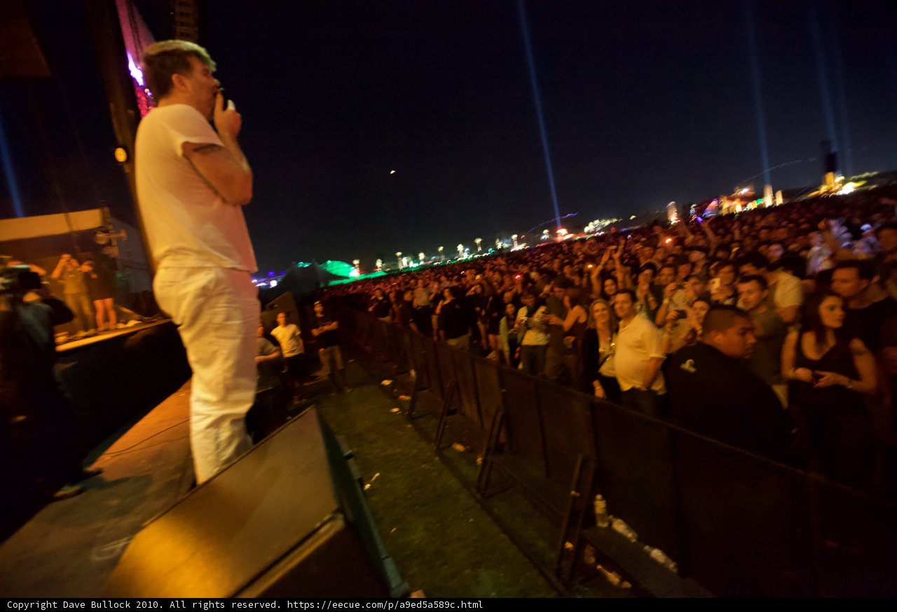 Rocking the Crowd Caption: This man takes center stage at the Cochella Friday 2010 concert, under the night sky, surrounded by a lively crowd of 18 people.