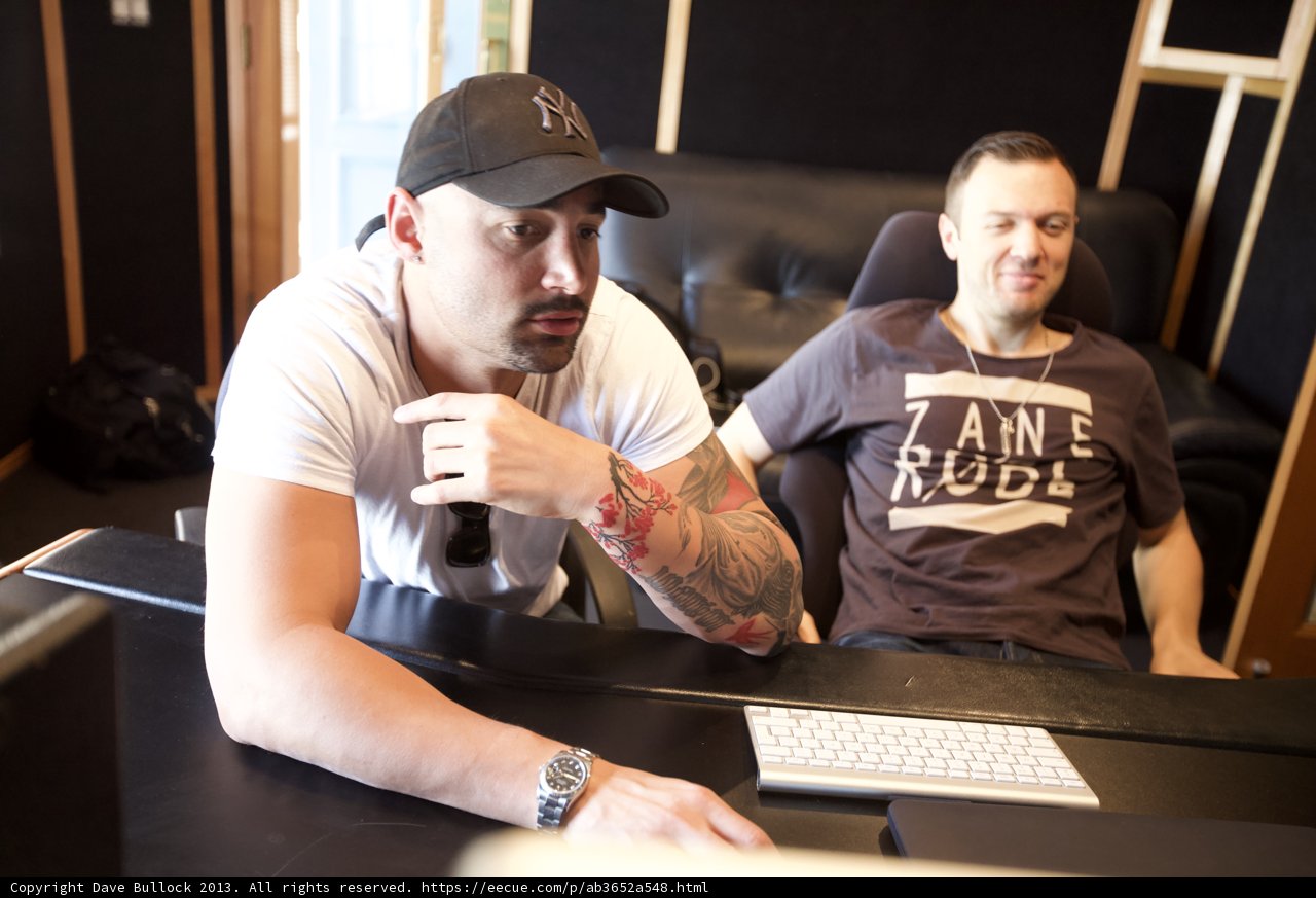 Recording Studio Session Caption: Chris Lake and a colleague sit at a desk in a recording studio in Altadena, California, surrounded by electronic equipment and music gear. Lake wears a baseball cap and a tattoo is visible on his arm, while his colleague works on a laptop.