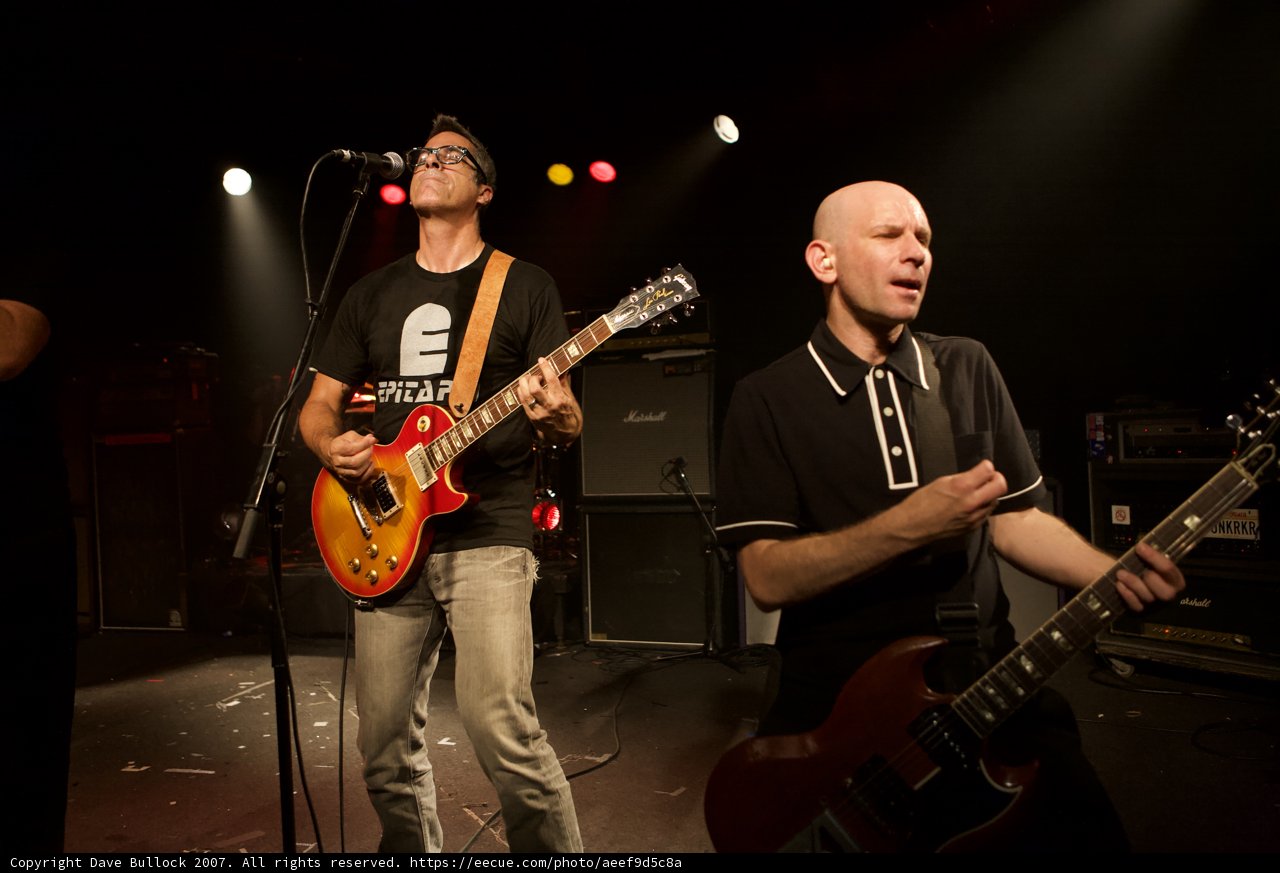 Two Guitarists Rocking the House Caption: Two talented men entertain a crowd with their guitars and microphones in a light-filled performance from the Bad Religion Glasshouse album in 2007.