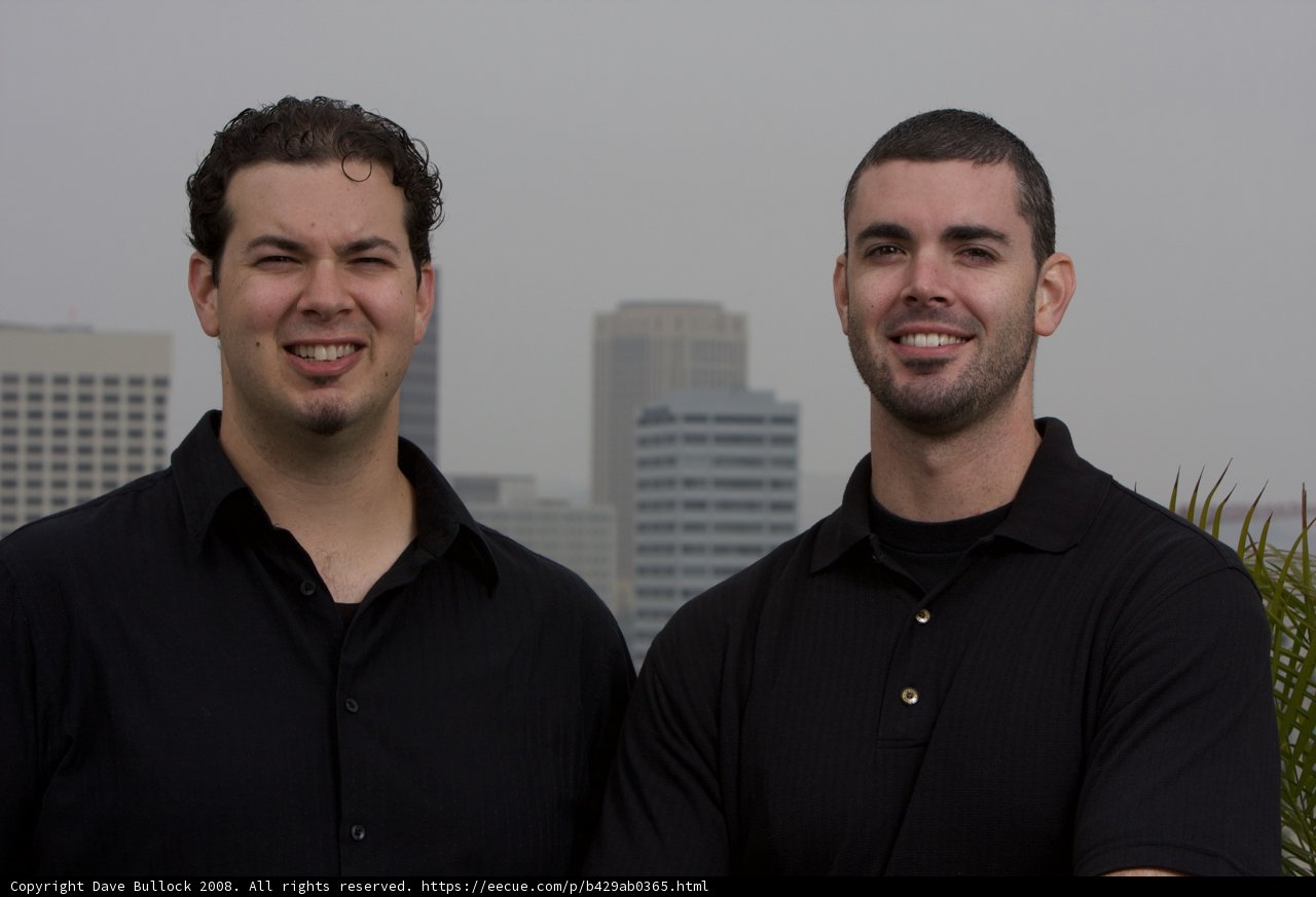 Two Smiling Men in Black Shirts Pose Next to Skyscraper