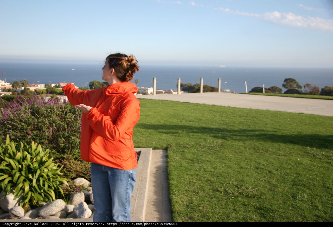 Woman in Orange Jacket Gesturing Toward Ocean View