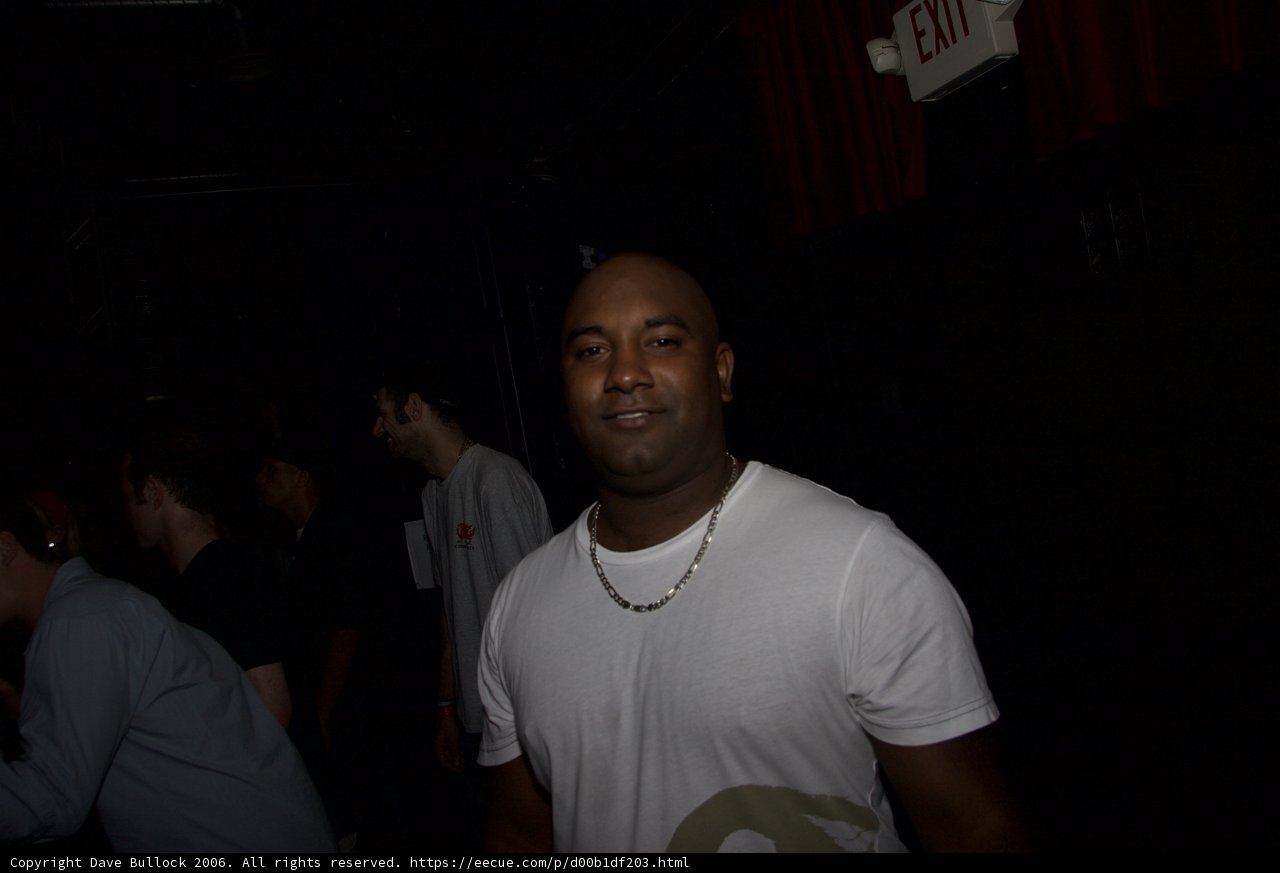 Commanding Attention Caption: A man stands confidently in front of a lively urban crowd at a night club wearing a T-Shirt and accessories, including a necklace and earbuds.