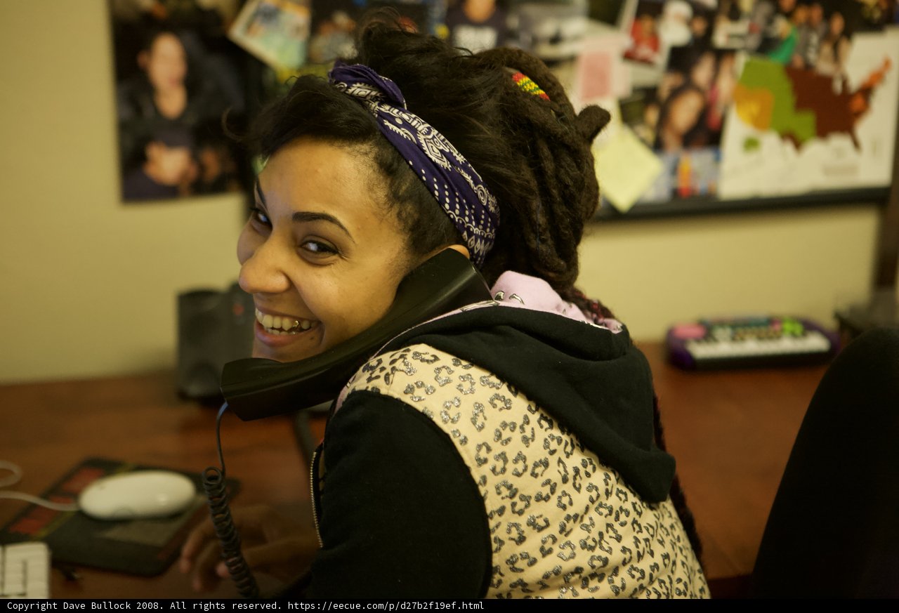 Dreadlocked Woman on the Phone Caption: Jodi G smiles while talking on the phone, surrounded by electronics and accessories at the APC office in 2008.