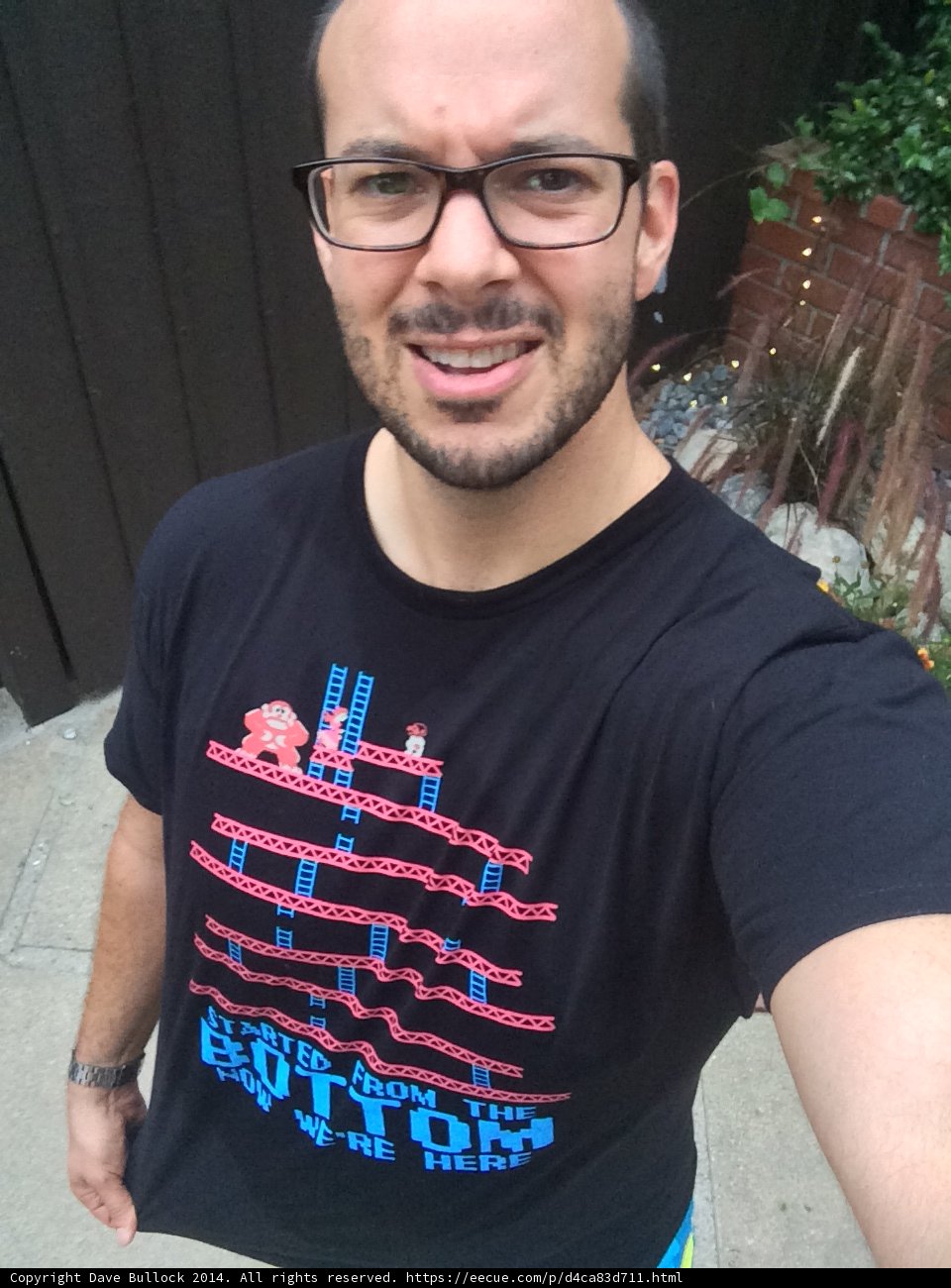 The Gamer Look Caption: Man posing in Altadena, California wearing a black T-shirt with a game design while accessorizing with eyeglasses and standing next to a decorative plant.