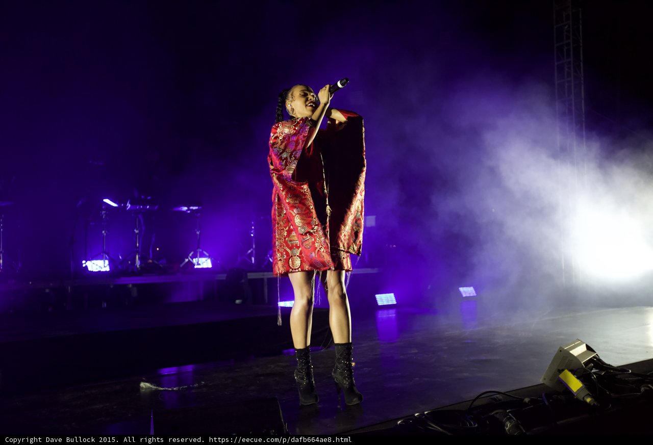 FKA twigs Full Body Standing Singing Microphone Raised Looking Up Legs Crossed Kimono Red Brocade Gold Patterns Black Boots Purple Lighting White Haze Band Truss LED Panels