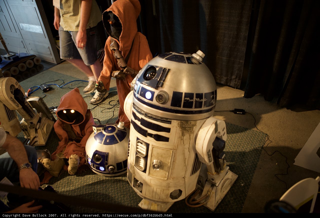 A Galactic Gathering Caption: Four people, including two babies, sit around a Star Wars R2D2 while wearing various clothing and headgear, including a helmet, and sporting two shoes and wristwatches.