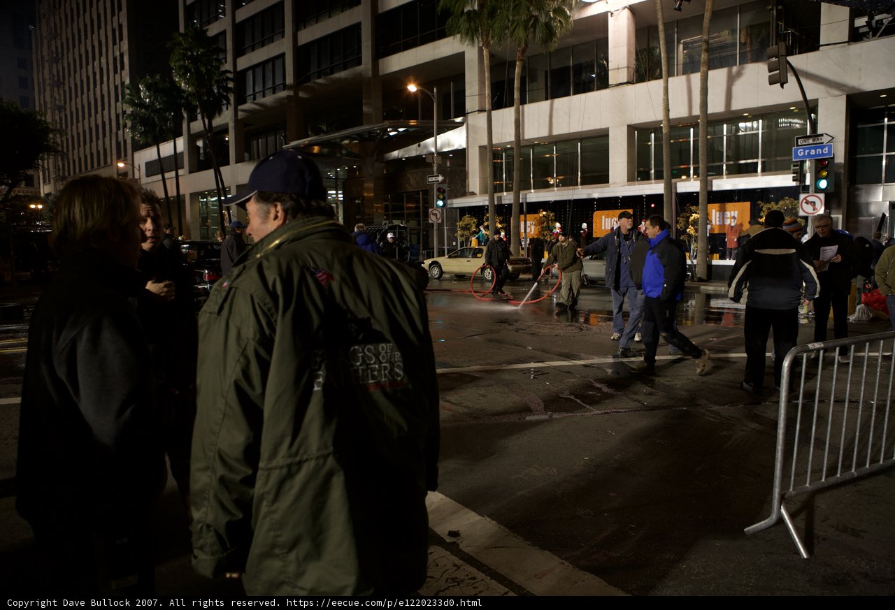Green Jacket in the City Caption: A man in a green coat walks down the busy sidewalk of a metropolis, surrounded by traffic lights, pedestrians, and towering palm trees.