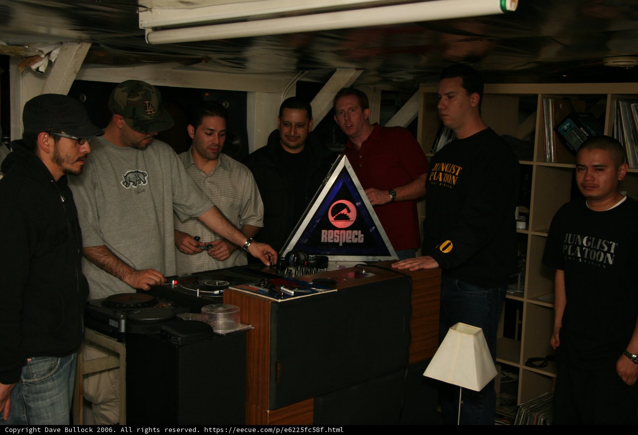 DJ Night Out Caption: Gil M, Justin F, Rob G, Clutch, and Cesar N stand around the DJ booth at the local pub, enjoying the music and the company. Two of them sport baseball hats and there are three wristwatches among the group. A lamp and an arcade game machine add to the atmosphere of the club.