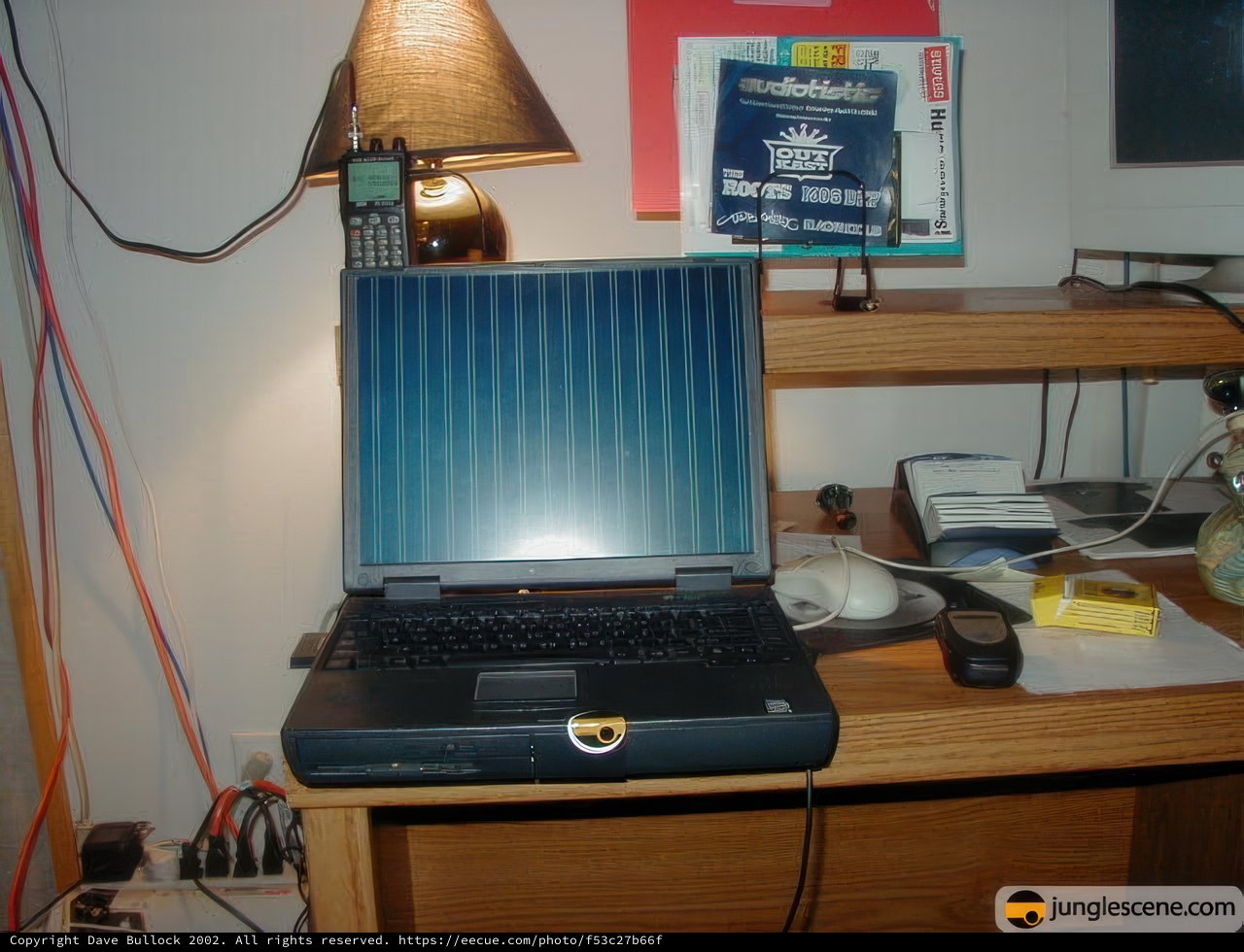 Working Hard Caption: A laptop computer on a desk covered in cords and surrounded by other electronics and furniture. A lamp shines down on the machine as it works diligently.